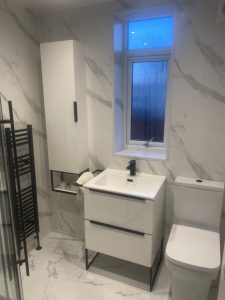 A modern bathroom featuring white marble walls with gray veins, a window with frosted glass, a white vanity with a black faucet and two drawers, and a white toilet. There is a tall white storage cabinet next to the vanity, and a black heated towel rail on the left side of the image. The floor is also covered in white marble tiles with gray veins, creating a cohesive and elegant design. The bathroom is well-lit and has a minimalist, contemporary aesthetic.