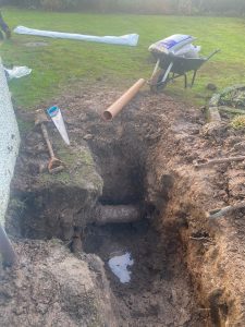 A large hole dug in a garden with exposed soil and some standing water at the bottom. A section of an old pipe is visible within the hole. Nearby, a new orange pipe and a handsaw are placed on the ground, and a wheelbarrow filled with bags of gravel or soil is positioned next to the excavation. The surrounding area features green grass and some bushes in the background. The scene indicates ongoing outdoor plumbing or drainage work.