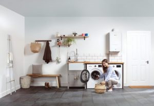 A woman kneels in a modern, well-organized laundry room, loading clothes into a front-loading washing machine. The room features a washing machine and dryer, a sink, shelves with various items, and a wall-mounted boiler. The floor is tiled in dark gray,​⬤