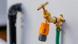 A close-up view of an outdoor brass faucet mounted on a white wall, with a yellow and gray hose connector attached to it. A green garden hose is connected to the faucet, secured with a metal clamp. The image is focused on the faucet and hose connection, with a blurred background that includes part of a black pipe. The scene is well-lit, showcasing the clean and functional setup for outdoor water use.